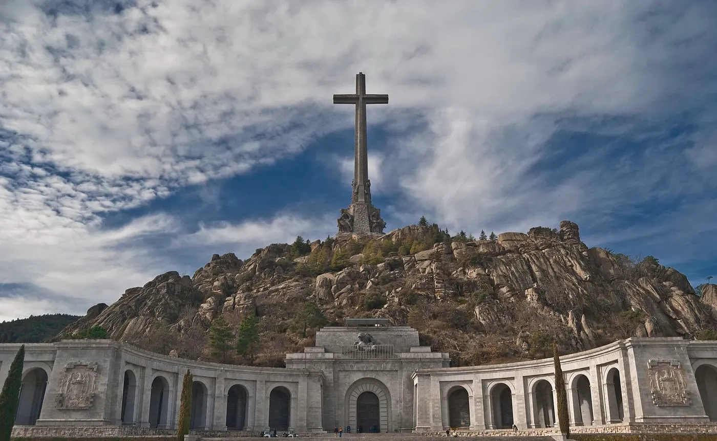 The tall stone cross on the granite hill above the Valle de Cuelgamuros, with the arcaded esplanade of the basilica below