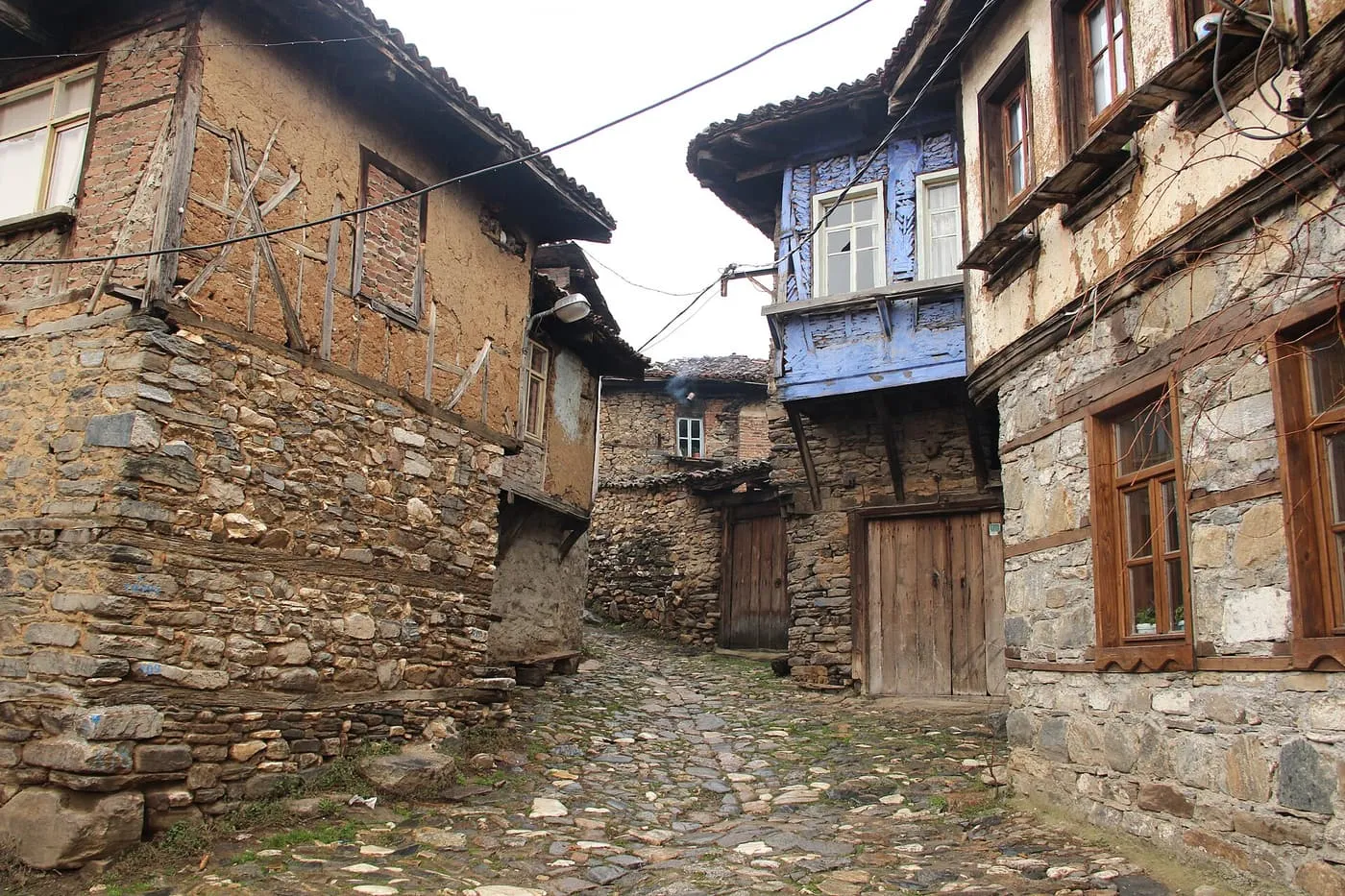 A cobbled lane in Cumalıkızık between old timber-framed houses with stone ground floors and wooden doors, one with a blue upper storey