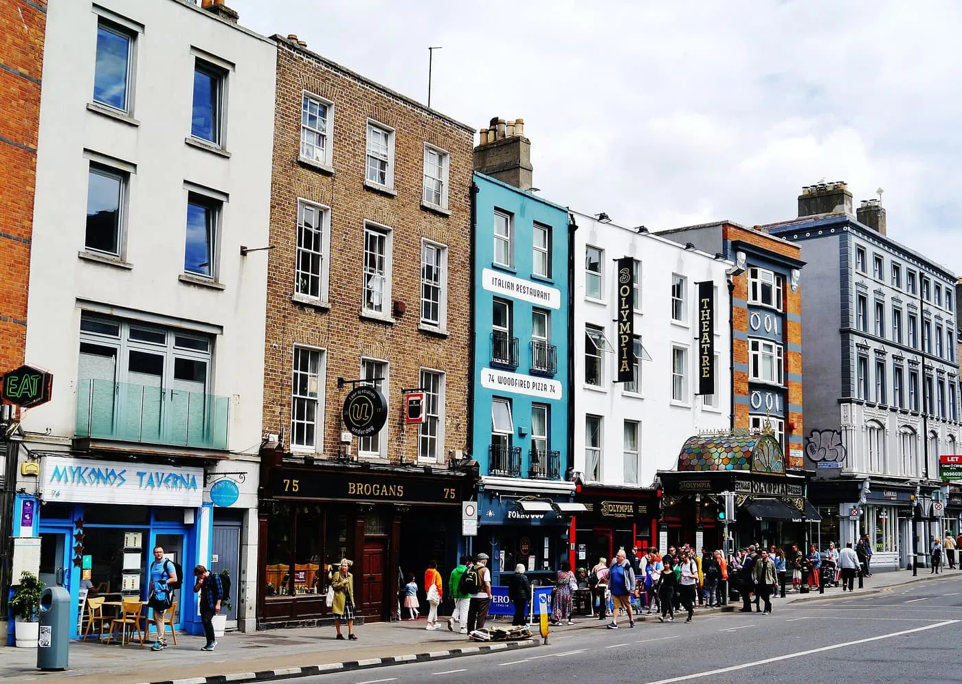 A row of painted shopfronts, pubs and the Olympia Theatre canopy along Dame Street, with pedestrians on the pavement
