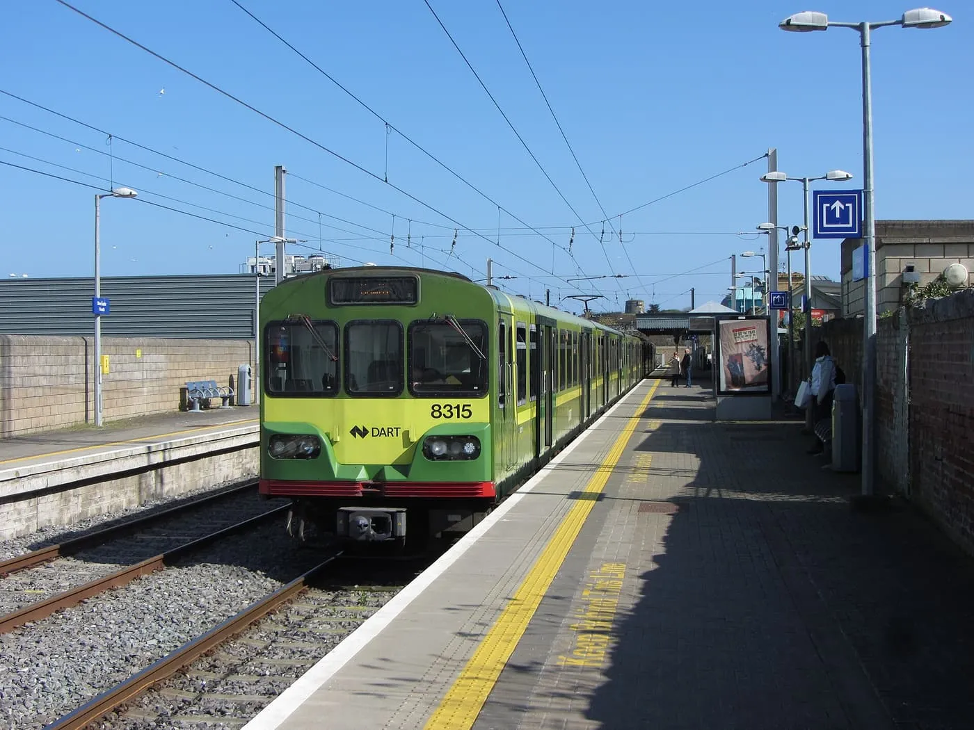A green and yellow DART train standing at a platform at Howth station under overhead wires
