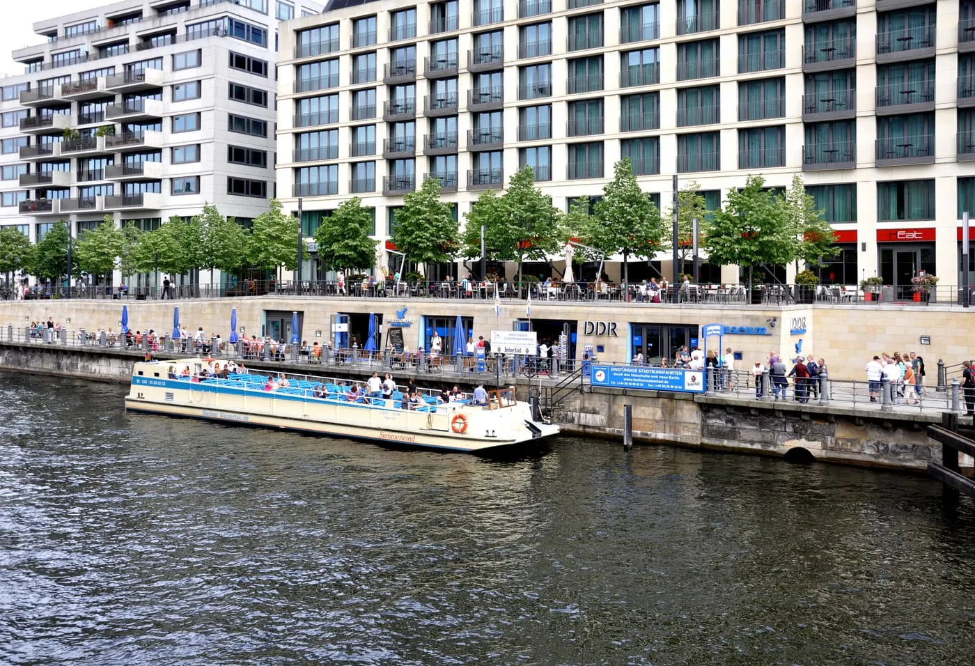 The DDR Museum on the Spree embankment, with its sign above the waterside entrance, a tour boat moored at the quay and apartment blocks behind