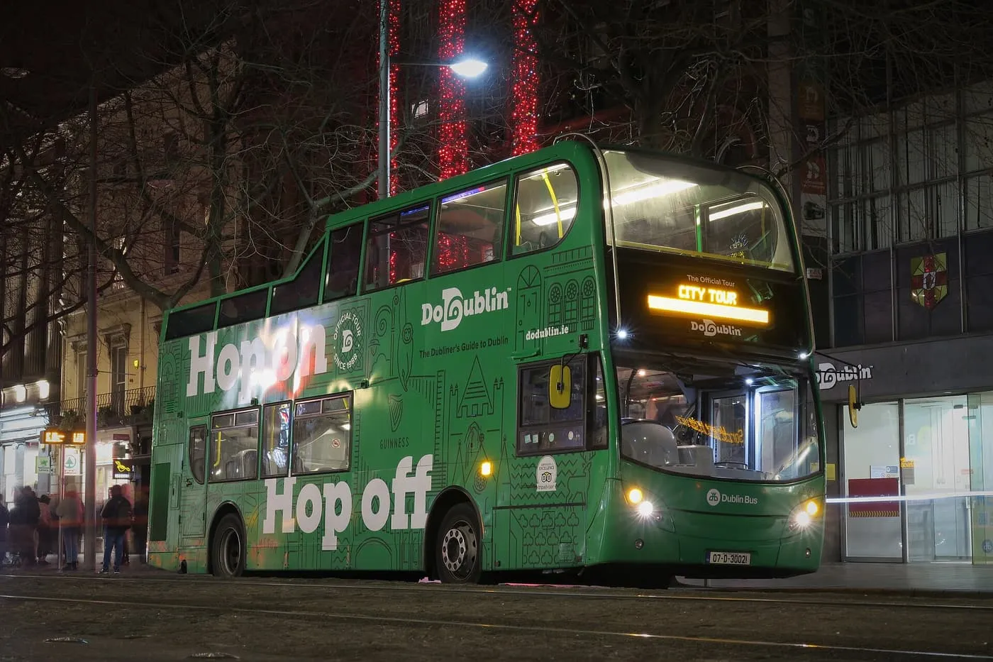 A green open-top DoDublin double-decker lit up at night with Hop on Hop off lettering along its side