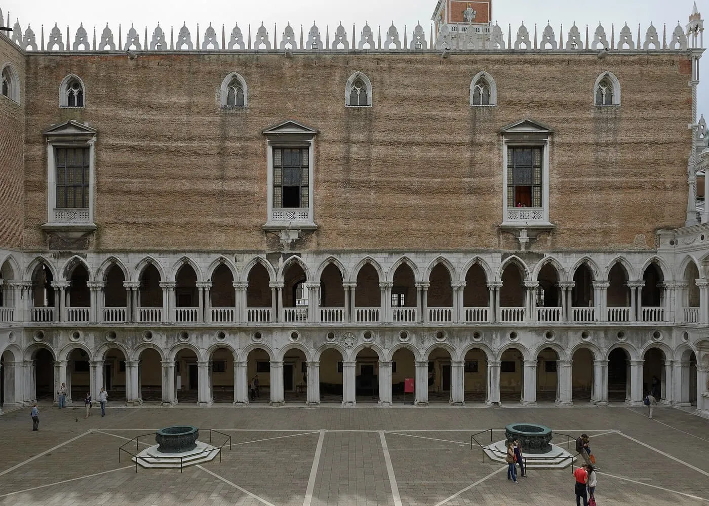 The brick west wall of the Doge's Palace courtyard above two storeys of white arcades, with a bronze wellhead on either side of the courtyard