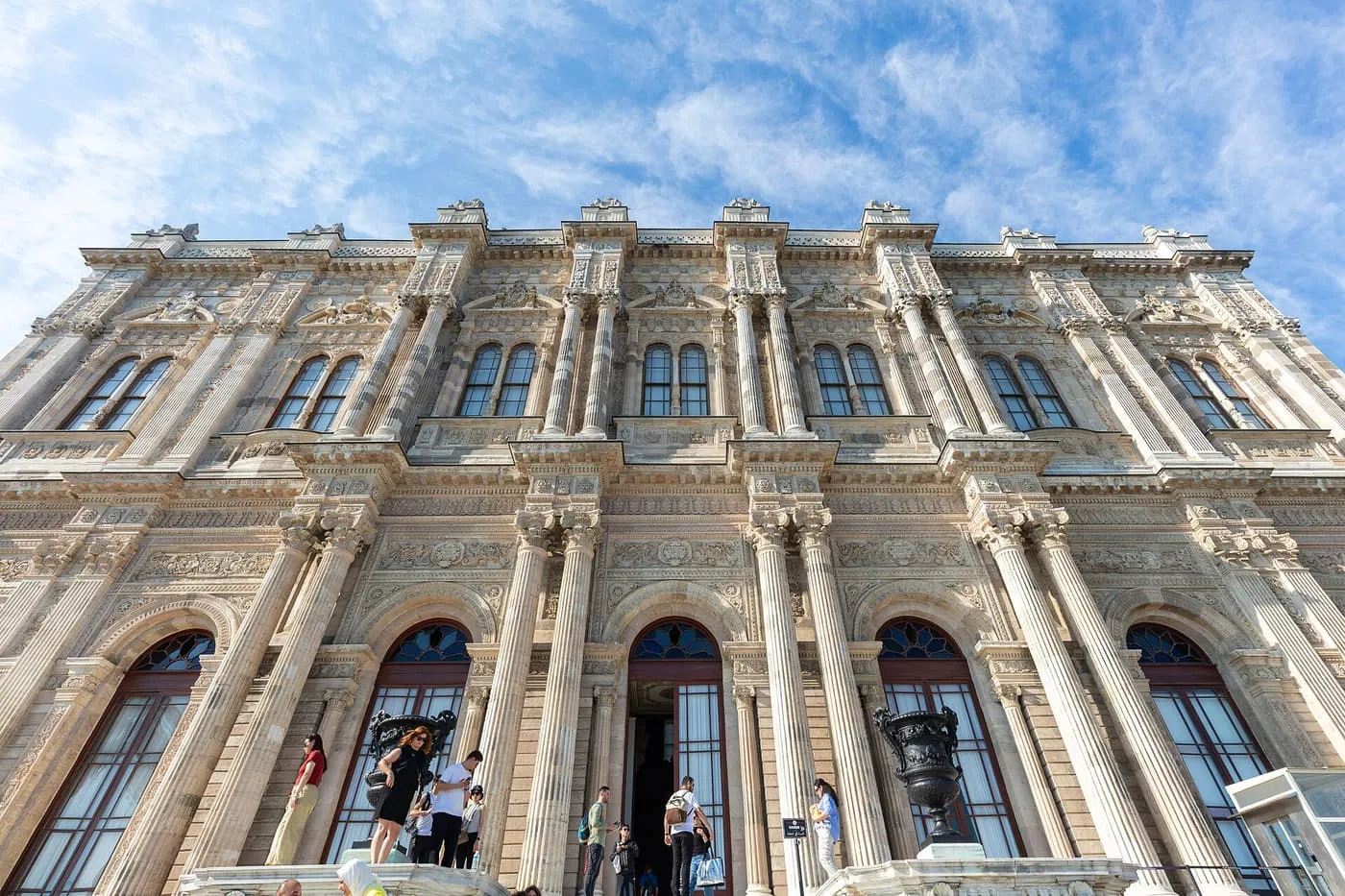 The carved stone facade of Dolmabahçe Palace, with tall arched windows and visitors at its doorways under a blue sky