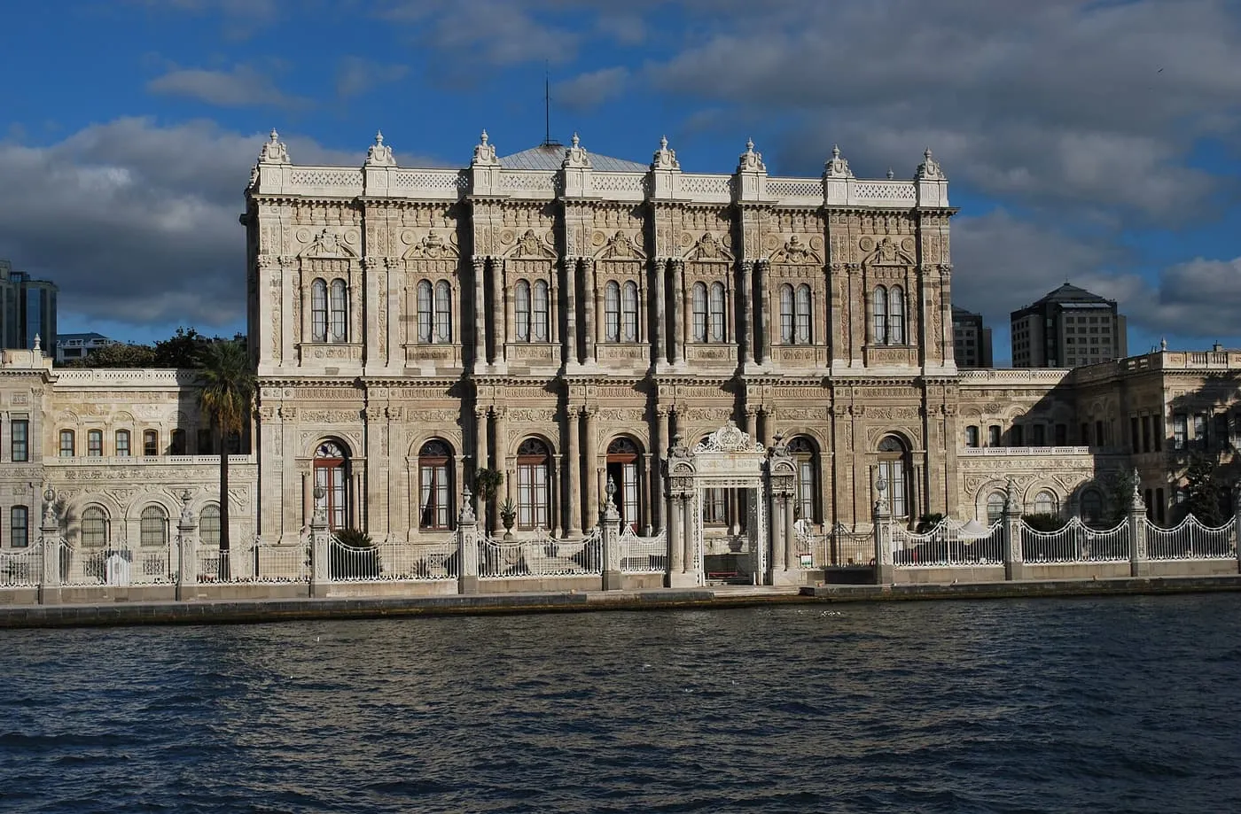 The long, ornately carved white façade of Dolmabahçe Palace with its gate and railings at the water's edge, seen from the Bosphorus