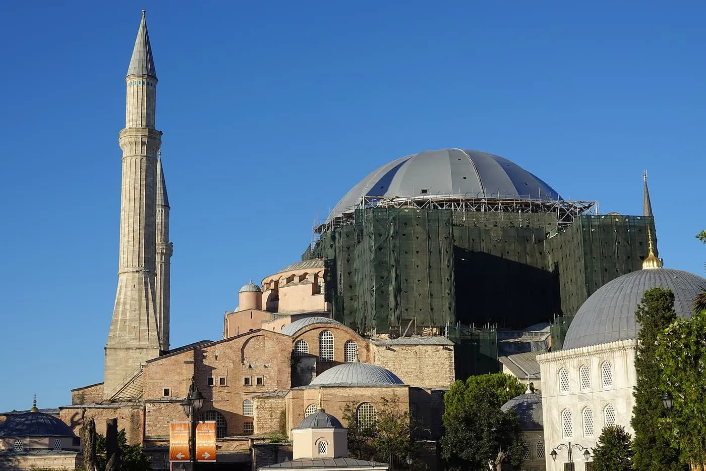 The grey main dome of Hagia Sophia ringed with scaffolding and green netting, between a minaret and a smaller domed building, under a blue sky