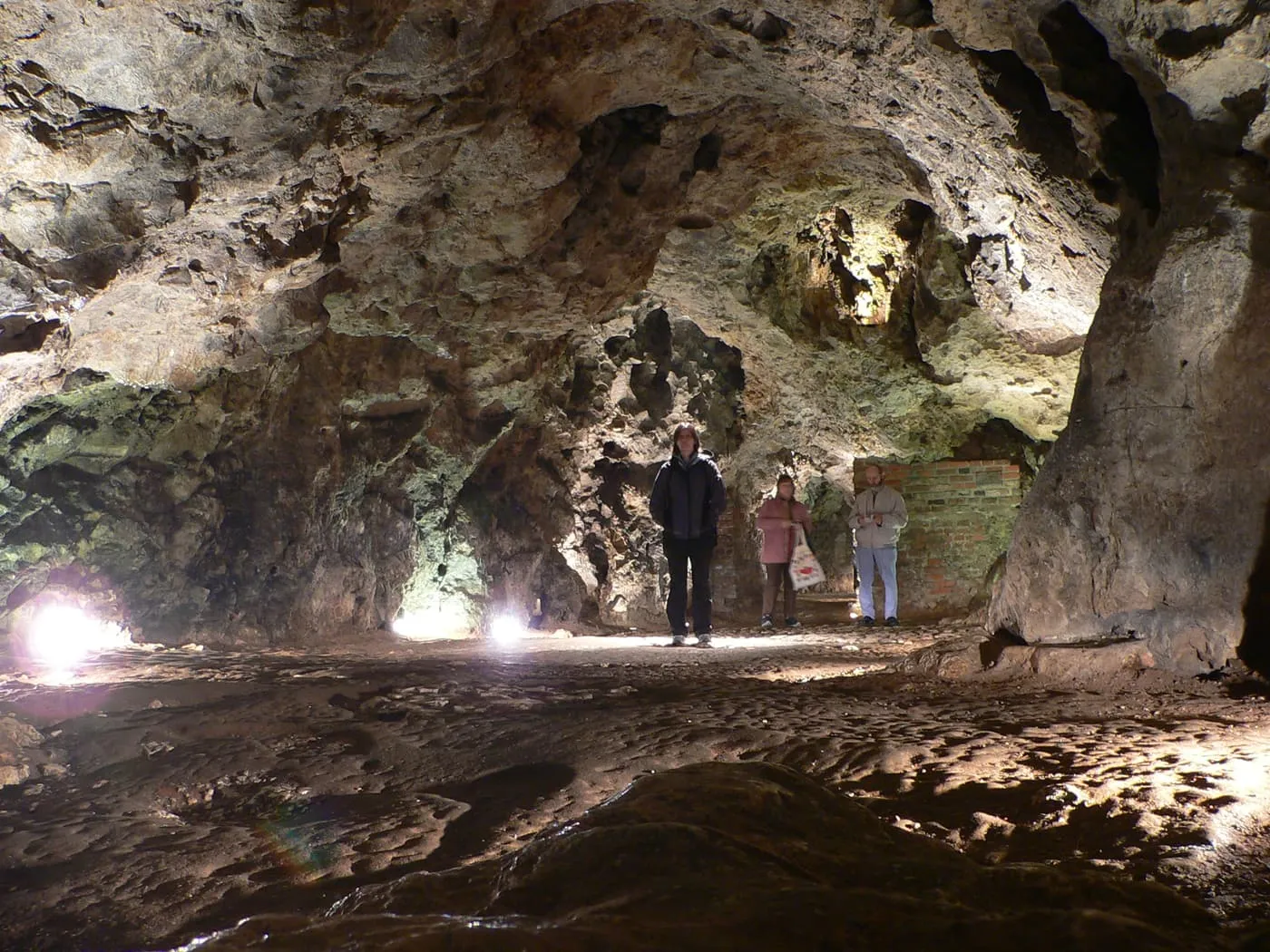 A limestone chamber of the Dragon's Den, lit from the floor, with three visitors standing at the far side