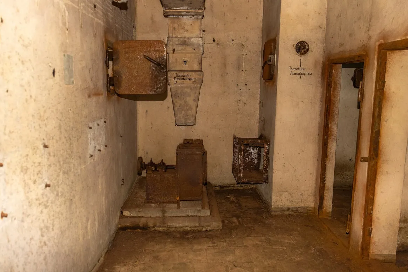 A bare concrete room with rusted ventilation equipment and a doorway, lit by torchlight