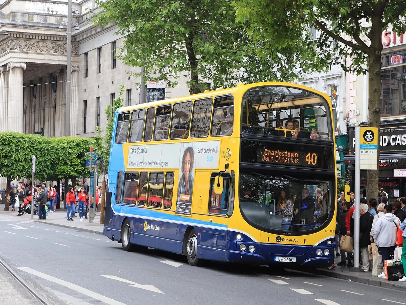 A yellow, blue and navy Dublin Bus double-decker on route 40 stopped at a kerb on O'Connell Street, with people waiting at the stop
