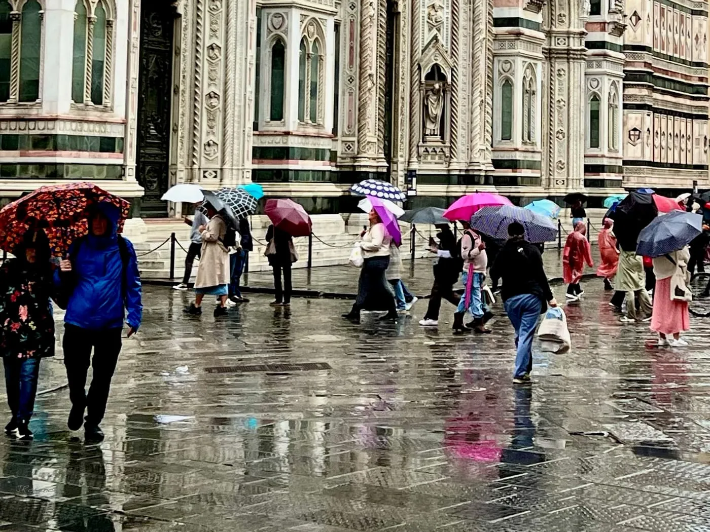 Visitors under umbrellas crossing the wet paving of Piazza del Duomo in front of the cathedral's marble facade