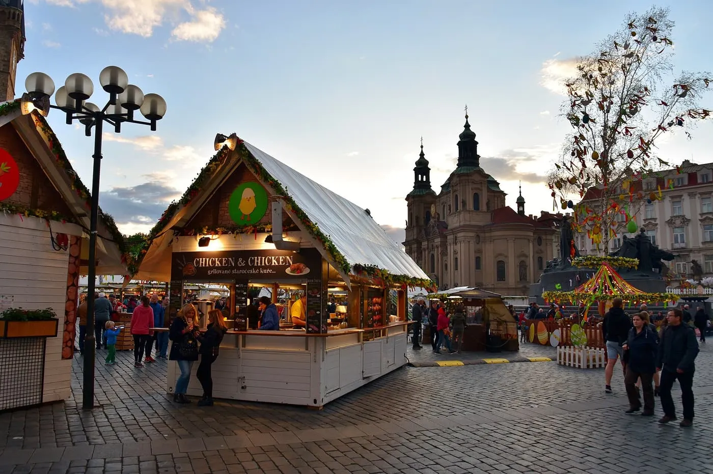 A stall with a green-and-yellow garland on its roof at an Easter market on Old Town Square, with the twin towers of St Nicholas Church and an egg-decorated tree behind