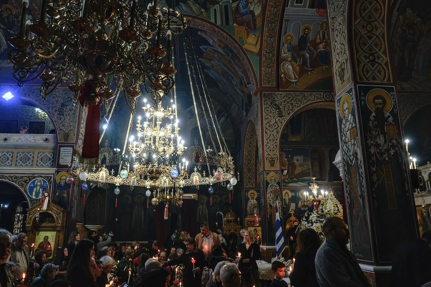 The packed interior of a painted Orthodox church during the Easter Vigil, with worshippers holding lit candles beneath a large brass chandelier and flower-decked stands at the side
