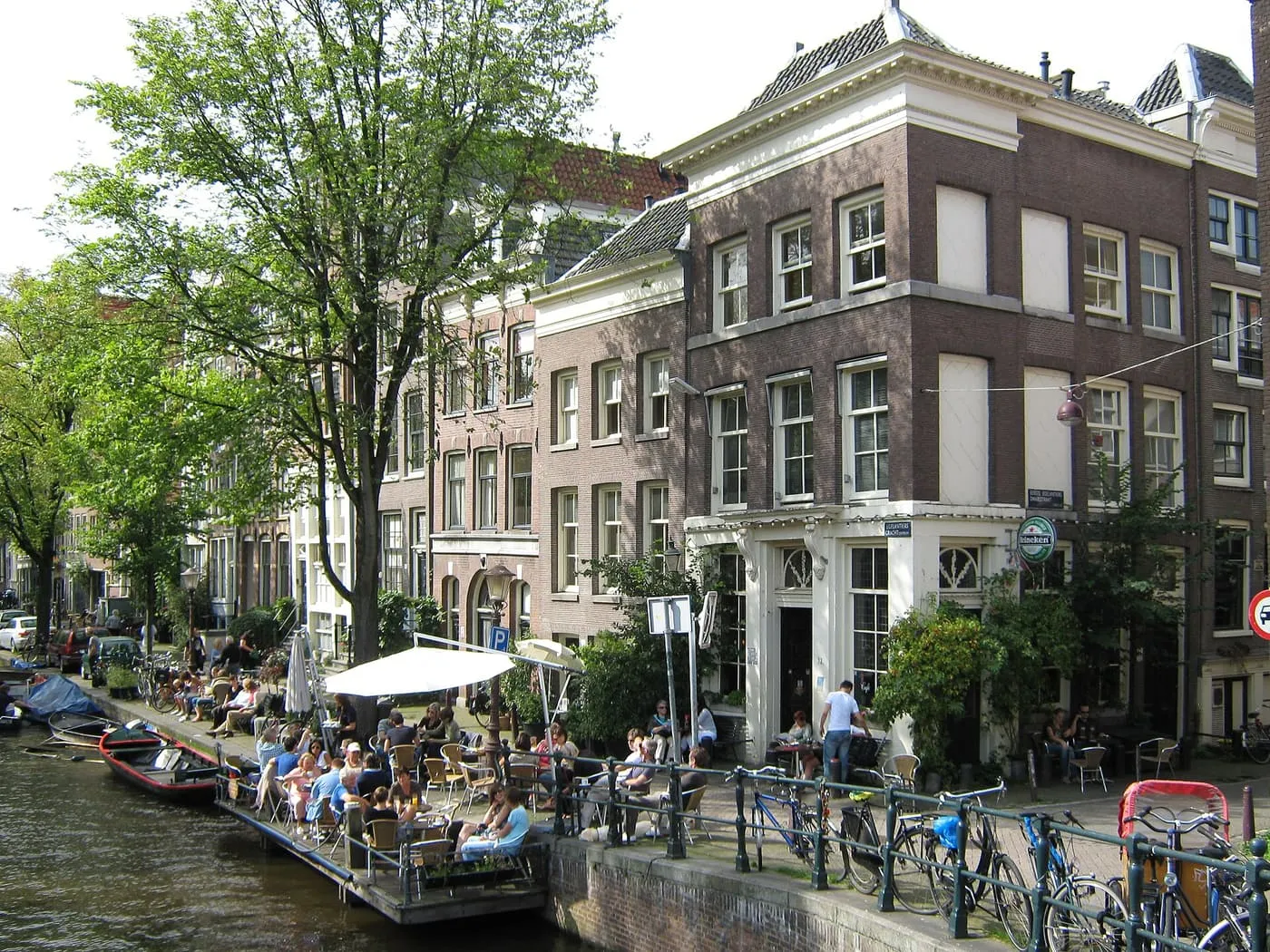Café tables under a white awning on a floating terrace beside the Egelantiersgracht, below a brown brick corner house with a Heineken sign