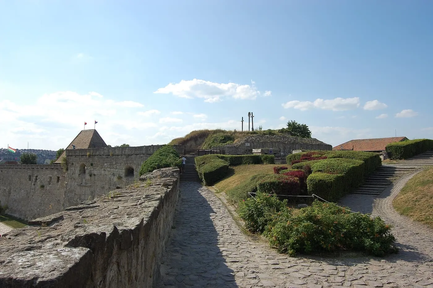 A cobbled path along the walls of Eger Castle, with a bastion roof and clipped hedges on the slope
