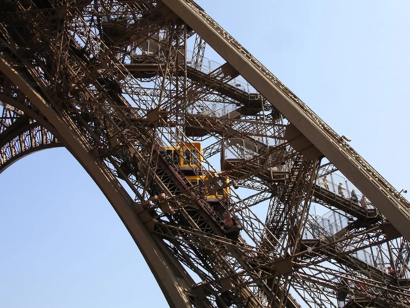 A yellow lift car climbing inside one of the Eiffel Tower's iron legs, beside stairways with people on them
