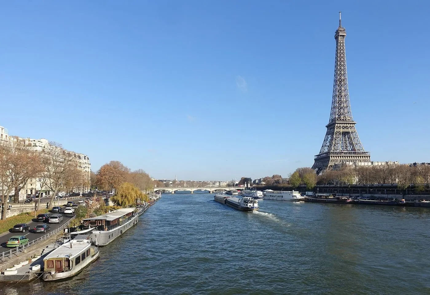 The Eiffel Tower beside the Seine on a clear day, with houseboats moored on the left bank of the river and a barge on the water