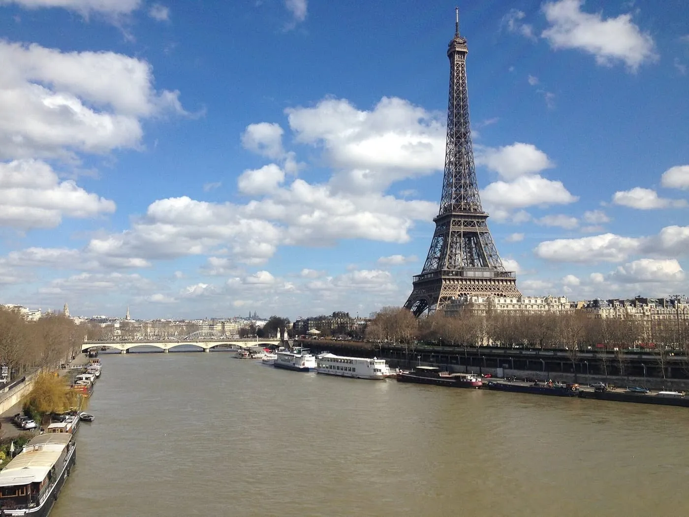 The Eiffel Tower above the trees and apartment buildings beside the Seine, with river boats moored along the quay under a blue sky
