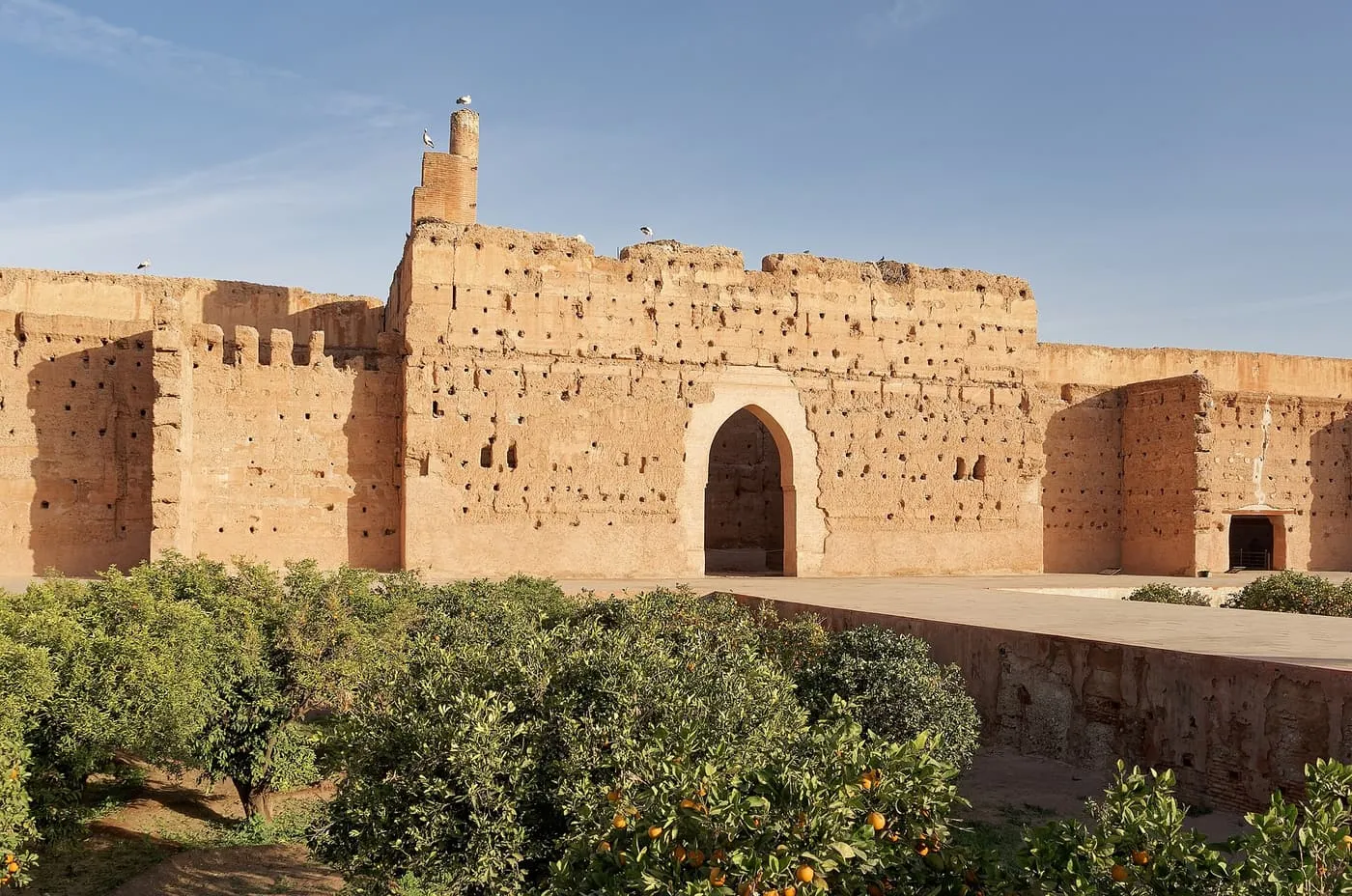 The ruined walls of El Badi Palace in warm stone, with an orange grove in front and storks standing on the top of the wall