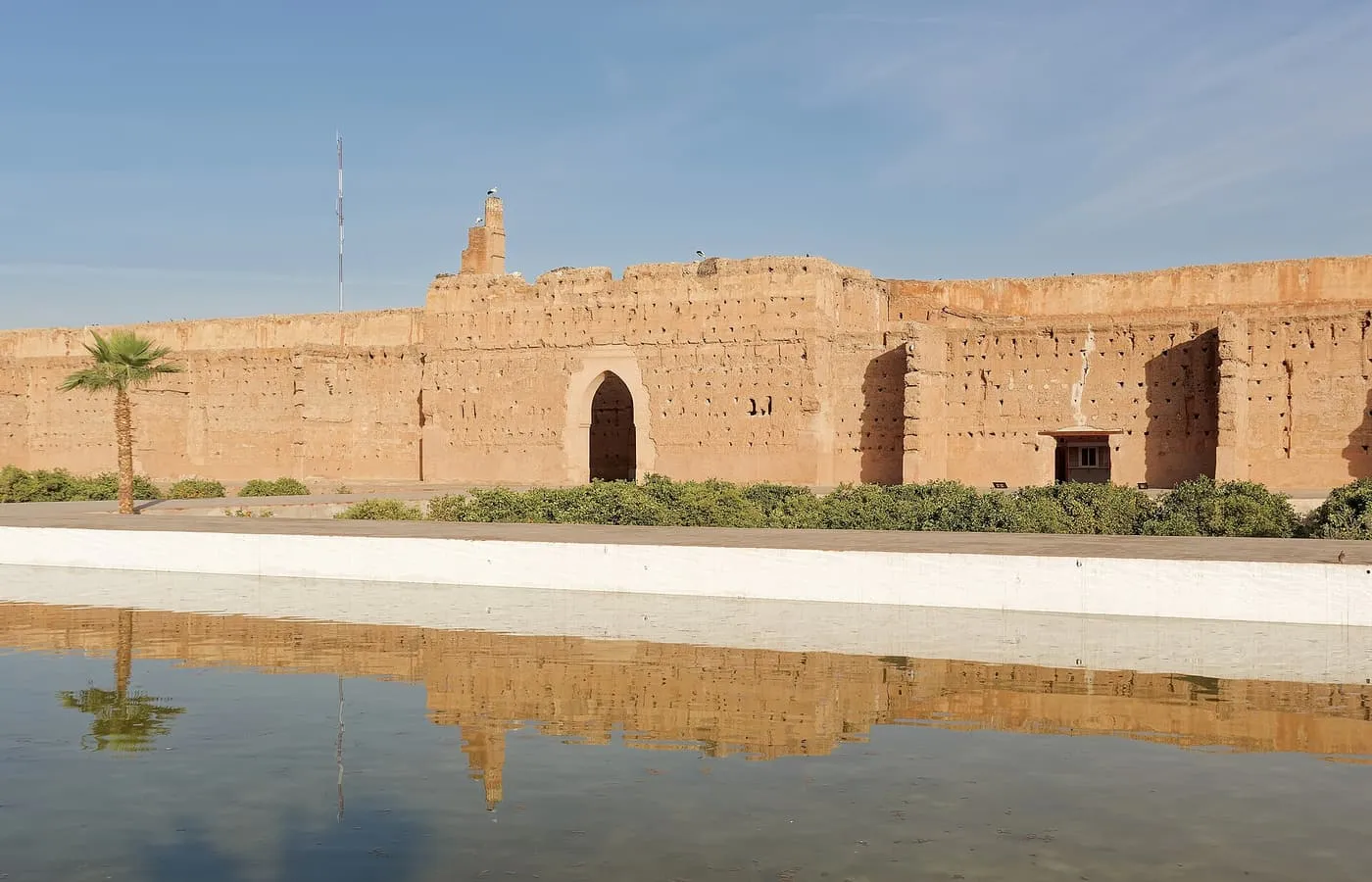 A long pool at El Badi Palace reflecting the ruined red-clay walls of the palace and a palm tree