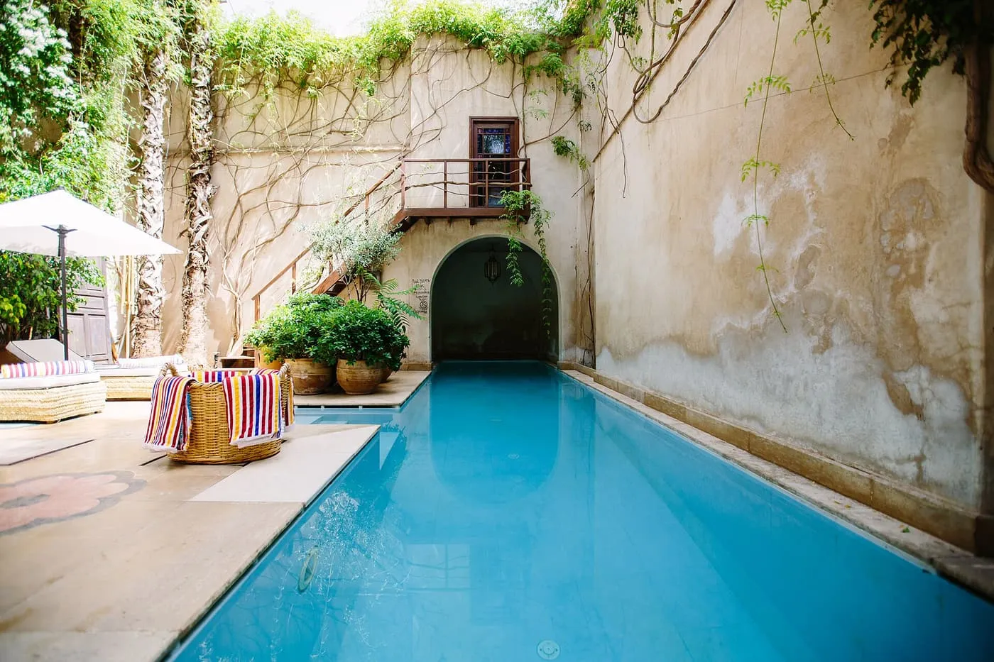 A long blue pool running into an arched passage between weathered walls hung with creepers, with a parasol and loungers at the near end