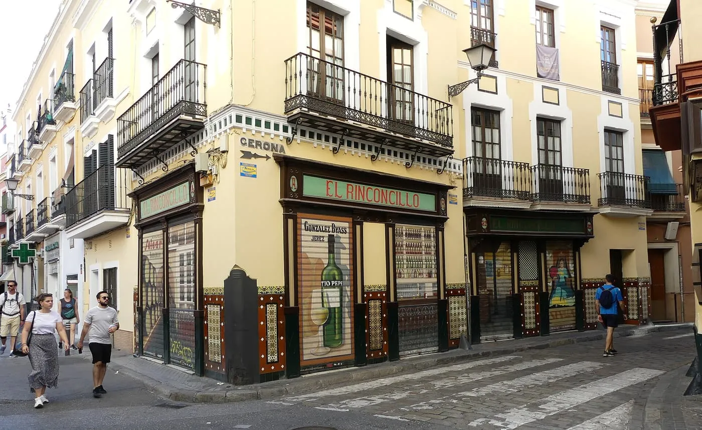 El Rinconcillo on a street corner, a cream building with iron balconies, a tiled base and a green sign over the door, with passers-by on the road