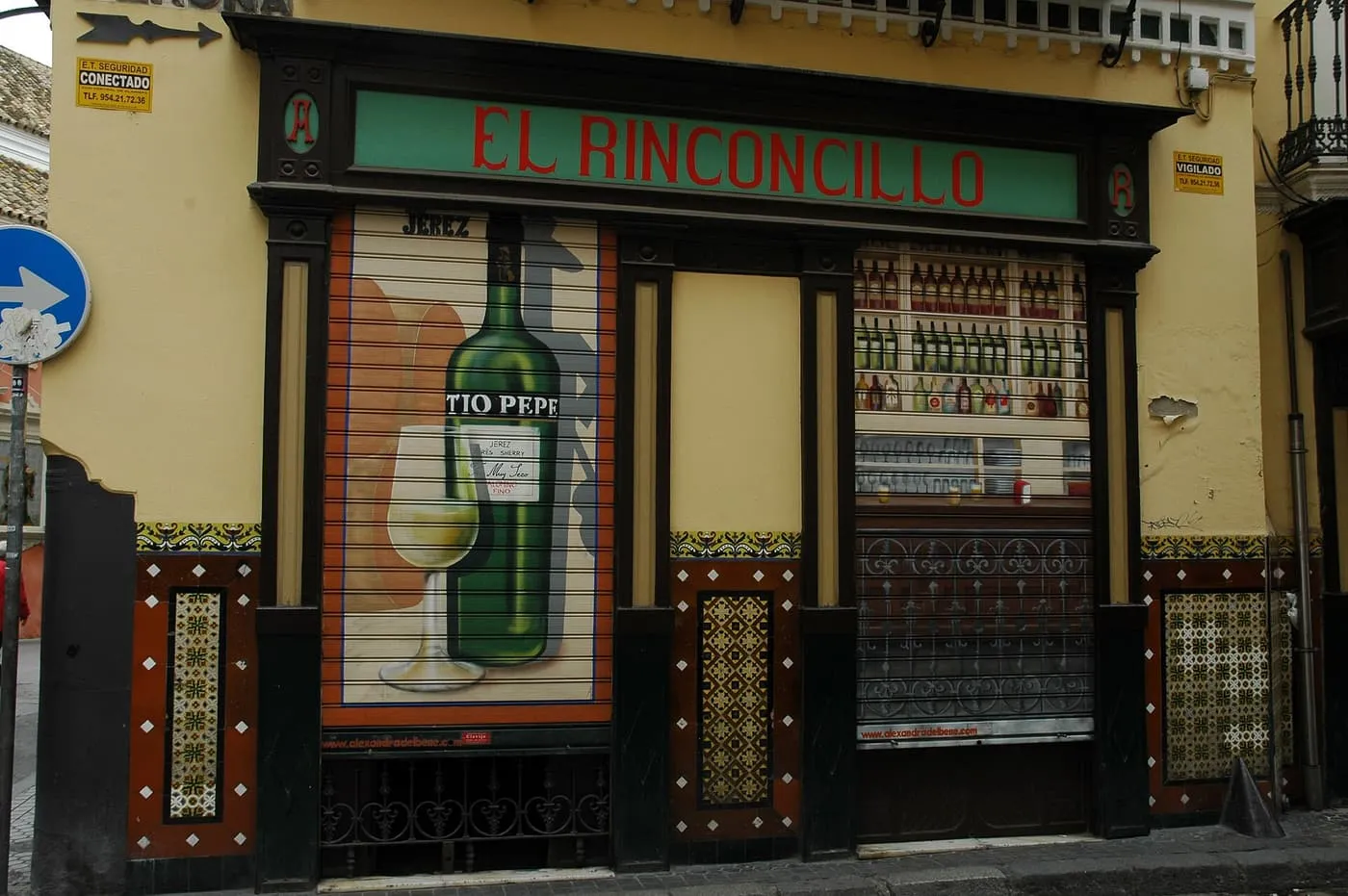 The front of El Rinconcillo, with a green and red sign, a Tío Pepe sherry mural on the shutter and tiles at street level
