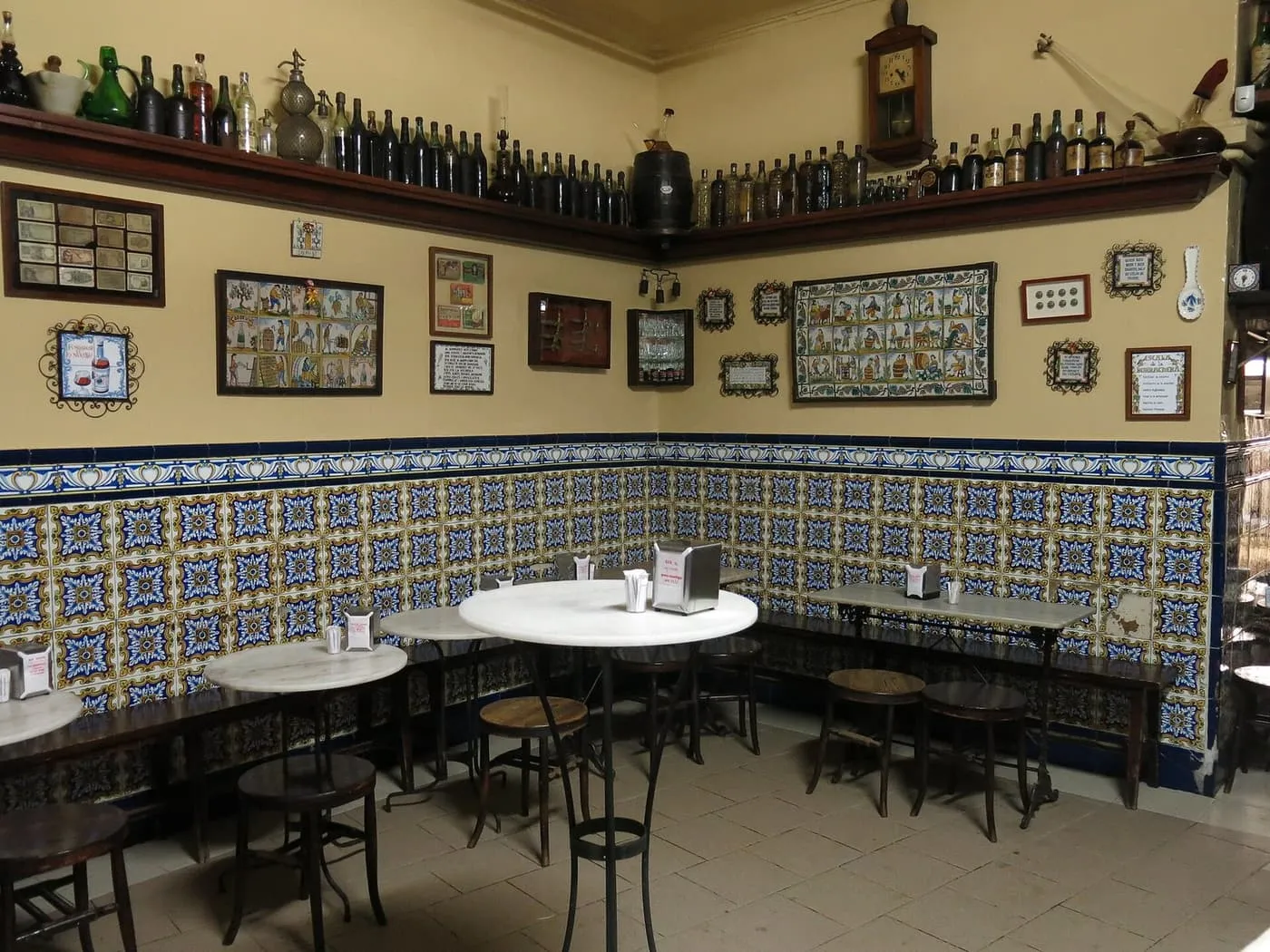 The tiled dining corner of El Xampanyet, with marble-topped tables, wooden stools and shelves of old bottles above blue and yellow wall tiles