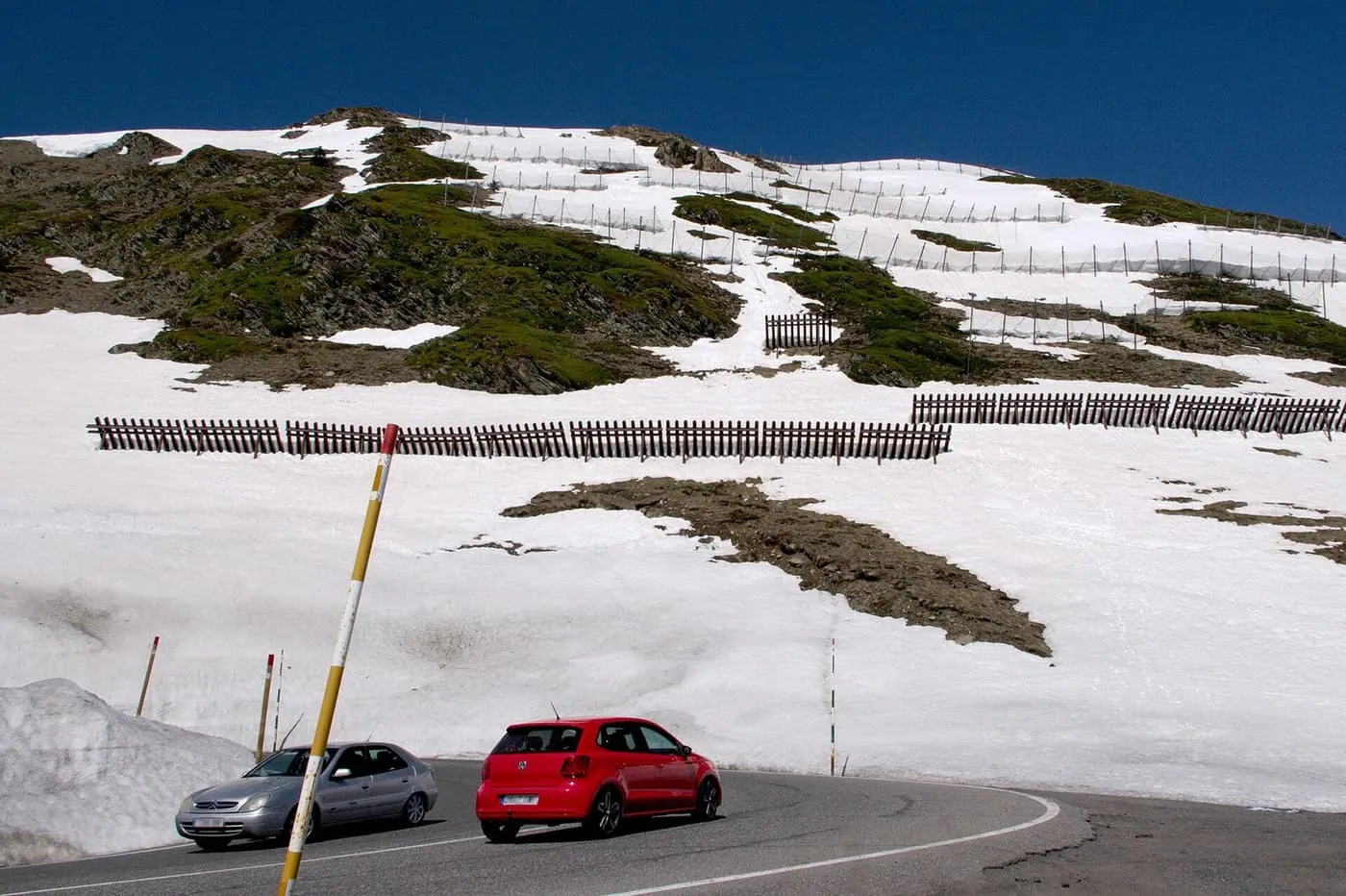 Two cars on a hairpin bend of the mountain road, with early summer snow and avalanche fences on the slope above
