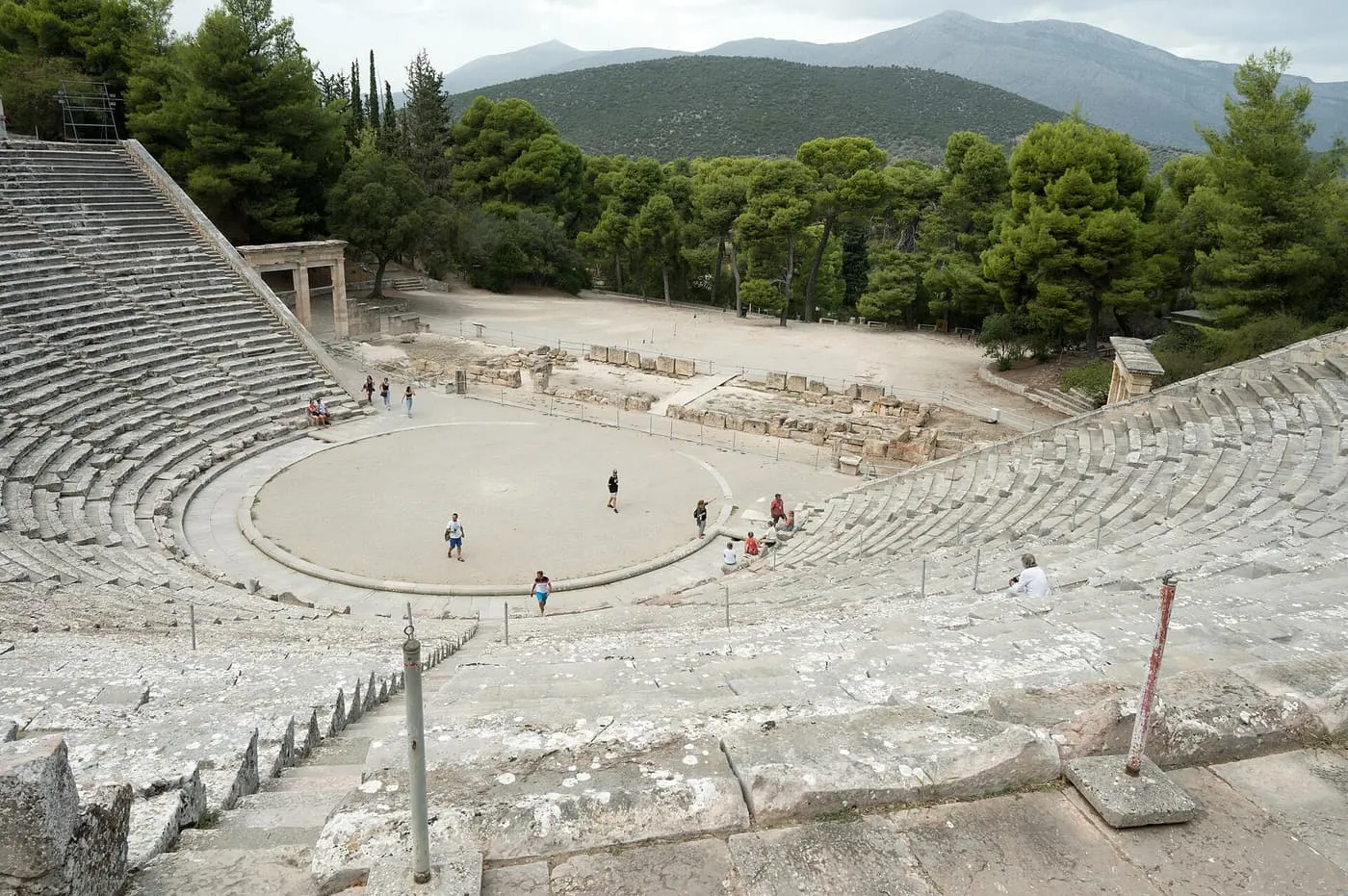 The curved stone seating of the ancient theatre at Epidaurus around a circular floor, with pine trees and wooded hills behind