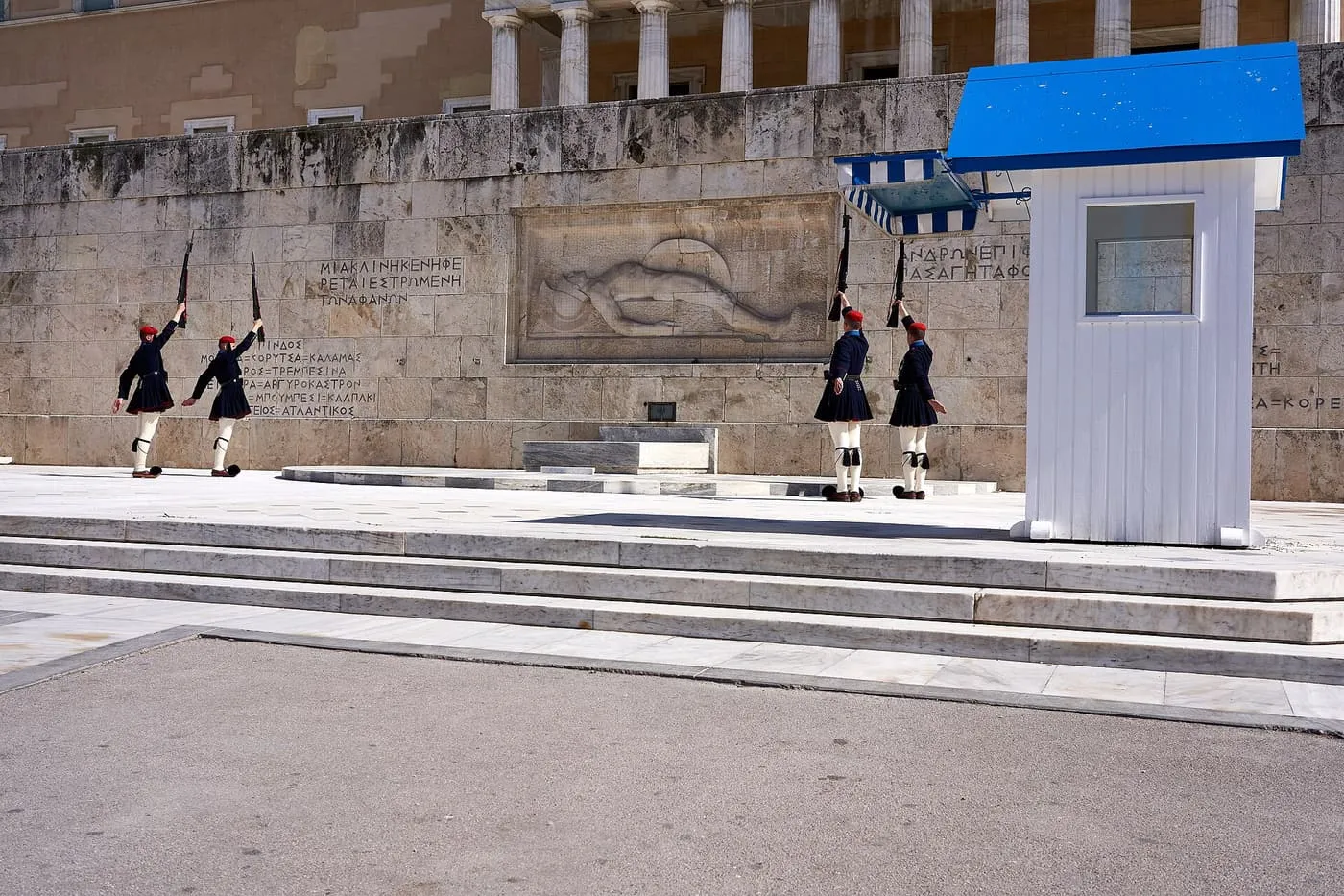 Four Evzones in dark winter uniform raising their rifles in front of the Tomb of the Unknown Soldier, with a white and blue sentry box and the Parliament building above