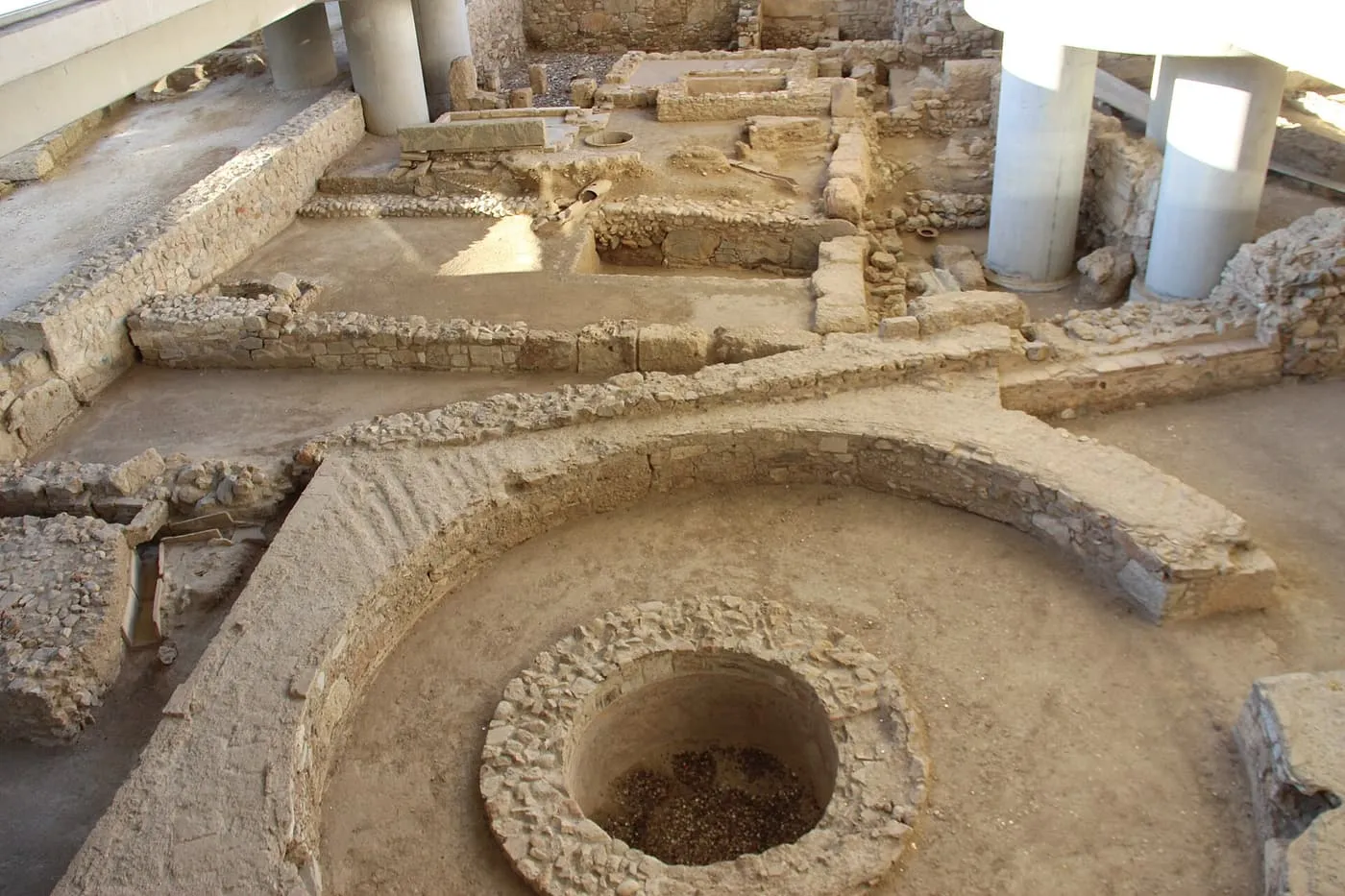 Excavated walls, floors and a round well of an ancient neighbourhood, with the concrete pillars of the museum standing among them
