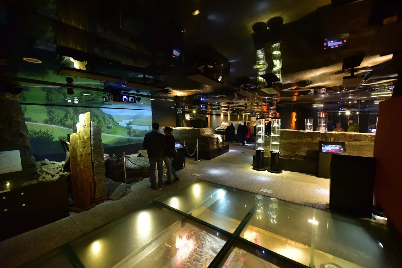 A dim exhibition hall of the Rynek Underground with a landscape projection, display cases and a glass floor over the excavations