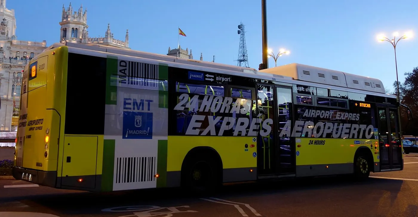 A yellow and green EMT Exprés Aeropuerto bus on the road beside the Palacio de Cibeles at dusk, with Atocha on its destination display