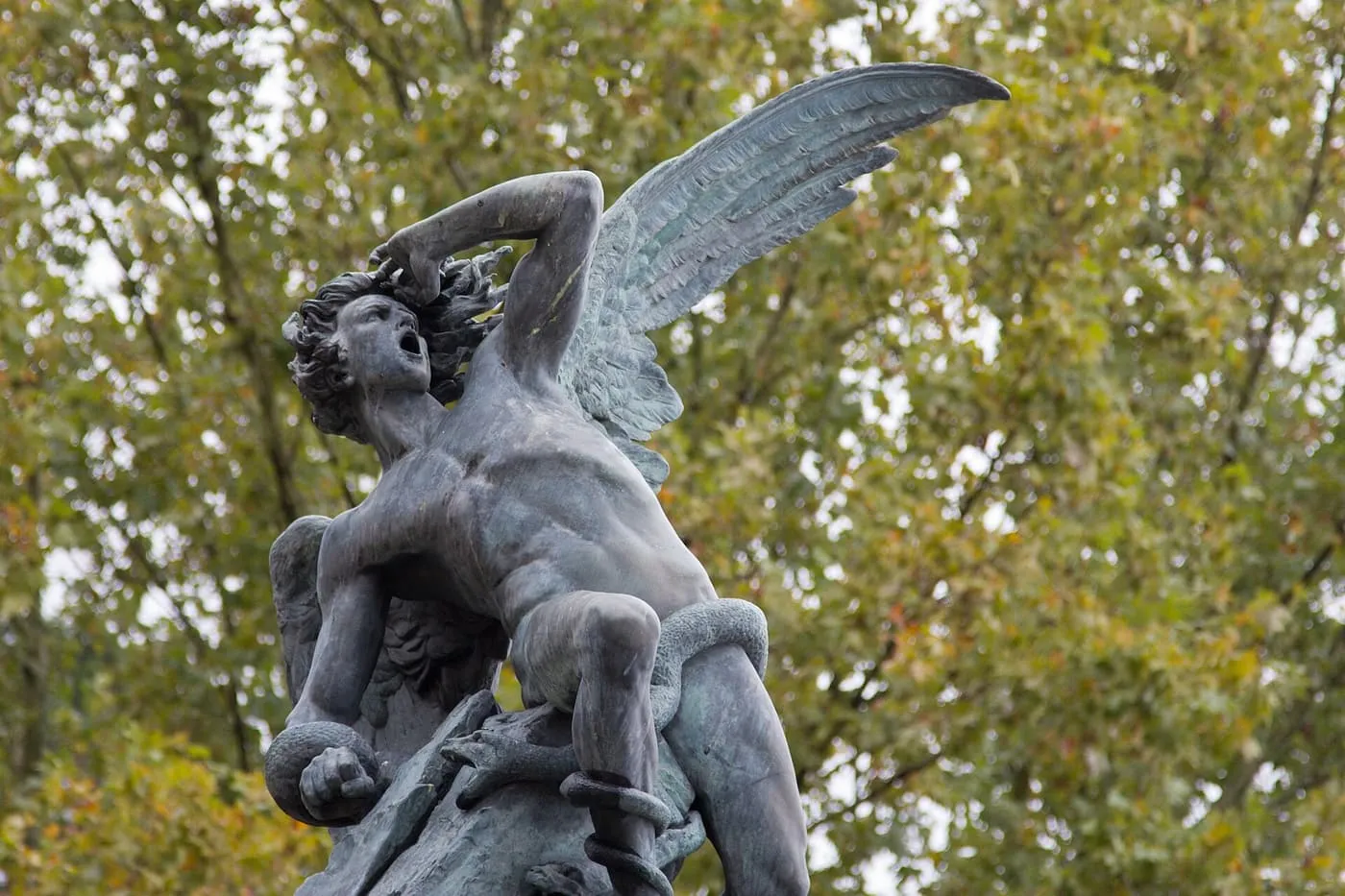 The bronze Fallen Angel statue in the Retiro, a winged figure twisted back with a snake round its leg, against autumn trees