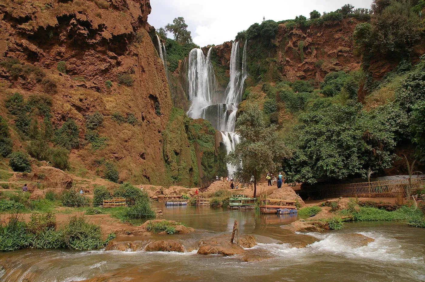The Ouzoud falls dropping in steps down a red cliff into a pool, with wooden rafts and visitors at the foot