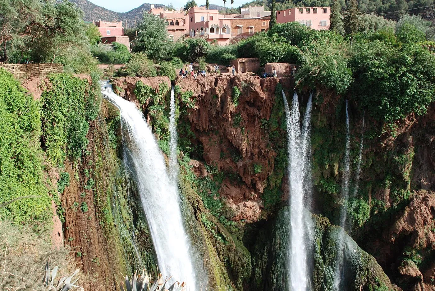 Two of the falls dropping past a red and green cliff, with visitors on the rim and pink buildings above