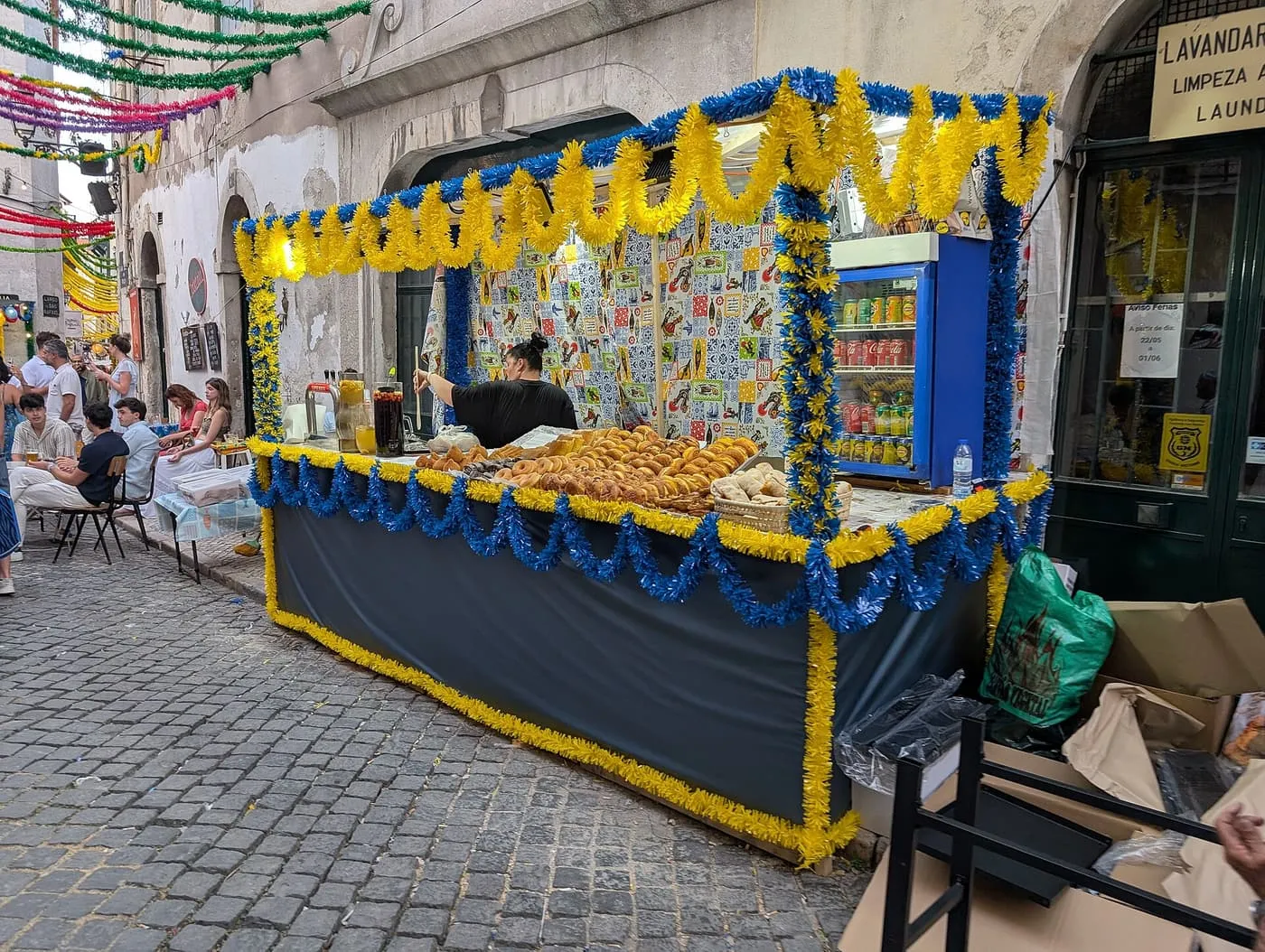A bakery stall dressed in yellow and blue streamers, with a counter of baked goods, on a cobbled Lisbon street
