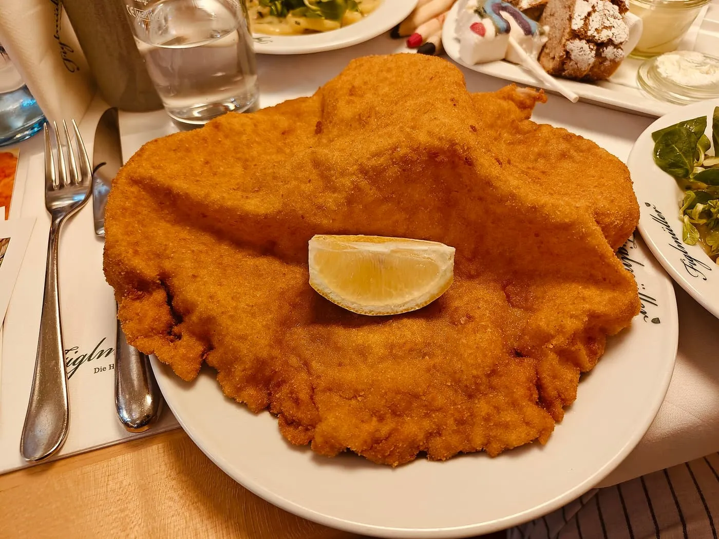 A pork schnitzel wider than its plate, breaded and fried golden, with a lemon wedge on top, on a table at Figlmüller