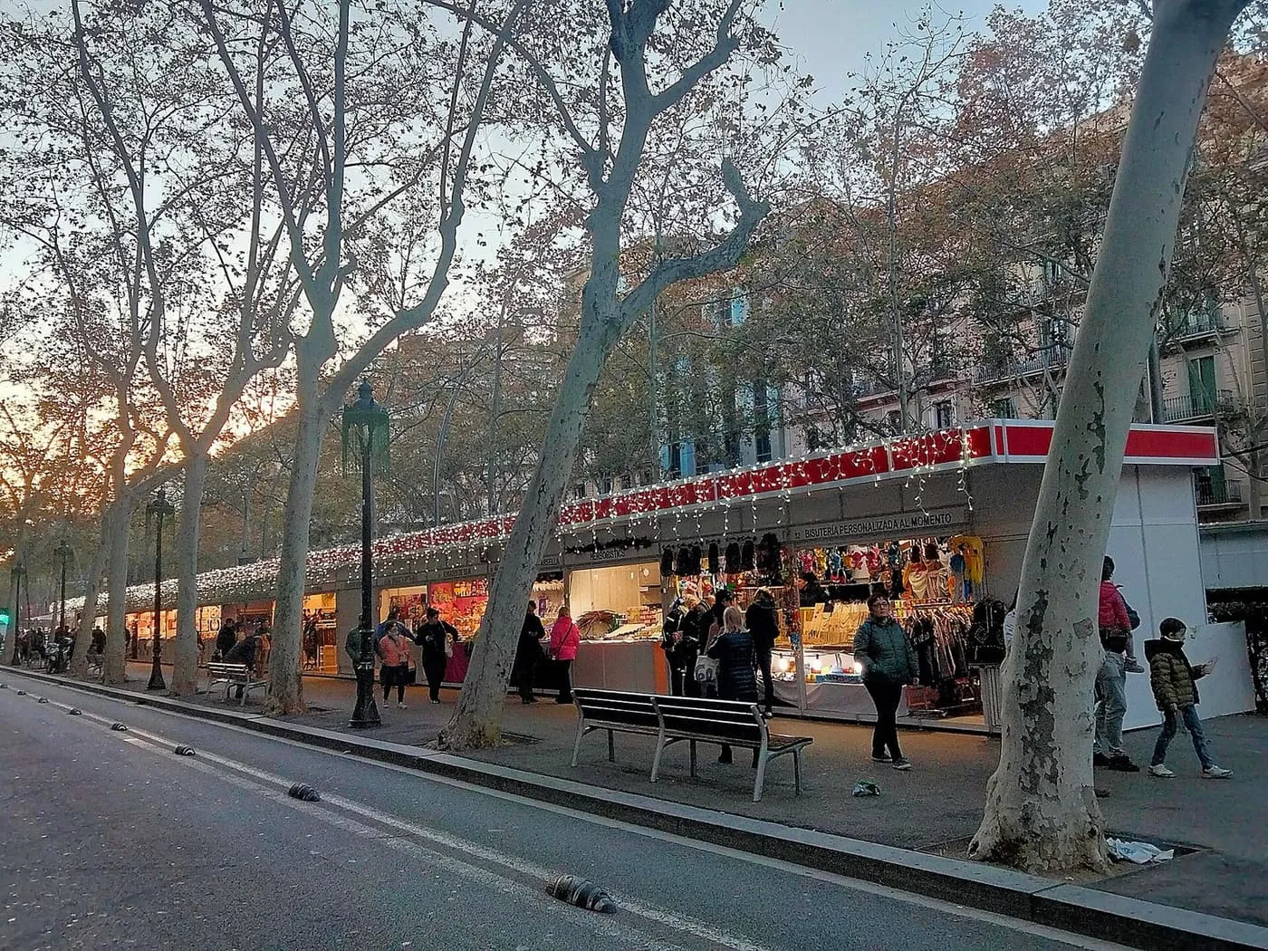 Fair stalls with a red-lit roofline along the Gran Via de les Corts Catalanes under bare plane trees at dusk