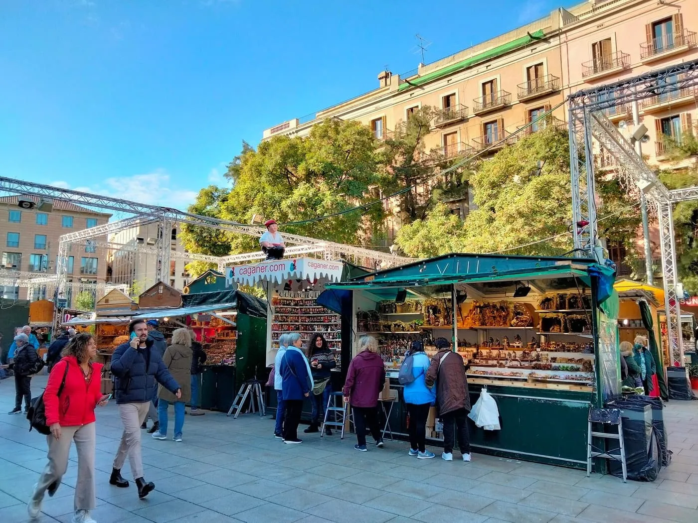 Wooden nativity-scene stalls of the Fira de Santa Llúcia on Avinguda de la Catedral, with shoppers, a caganer figure on a stall roof and a blue winter sky
