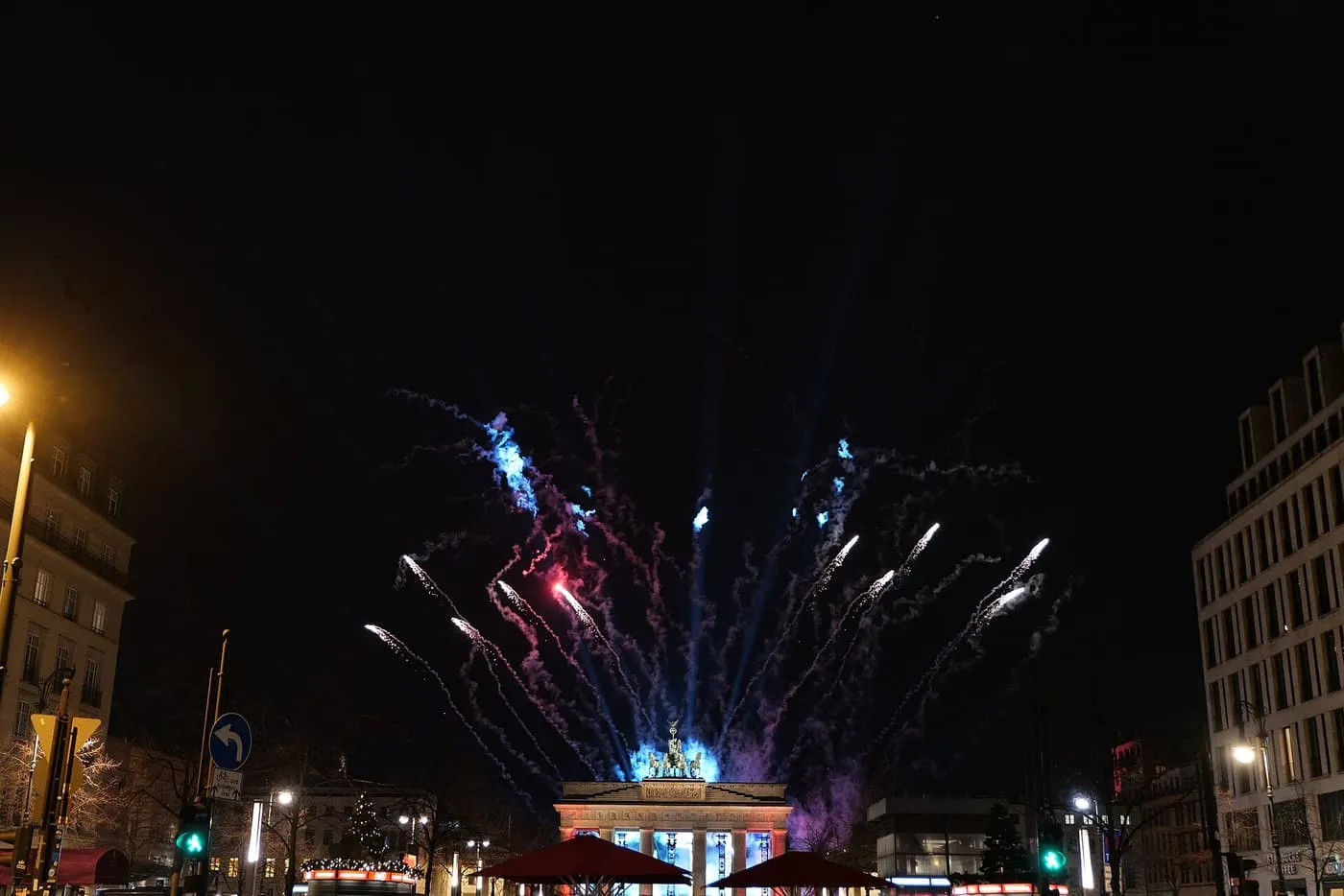 Blue and white firework trails rising above the Brandenburg Gate at night, seen along Unter den Linden
