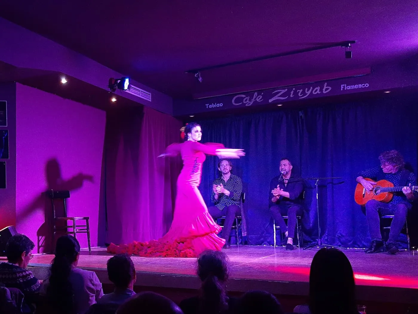 A flamenco dancer in a long red dress turning on a small stage lit in magenta, with a singer, a clapping musician and a guitarist seated behind her and the backs of the audience in front