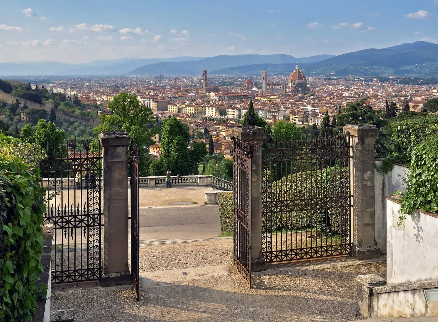 Florence seen between iron gates at San Miniato al Monte, with the Duomo dome and a tower among the roofs and blue hills beyond