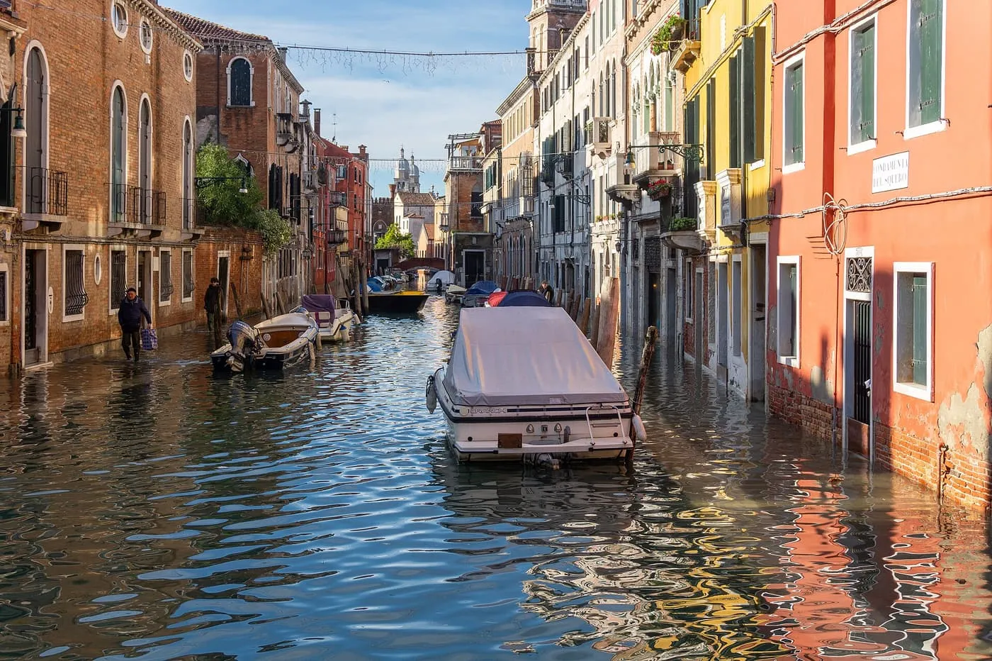 A flooded canal-side street with water up to the doorsteps, a covered boat in the channel and a man wading