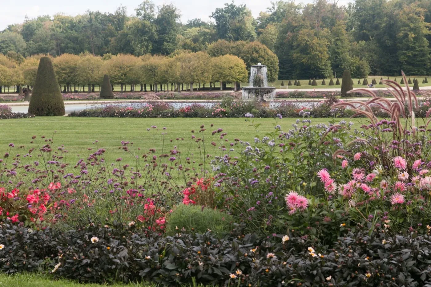 A fountain in a pool beyond a lawn and flower beds of dahlias and verbena, with clipped yews and lines of trees behind