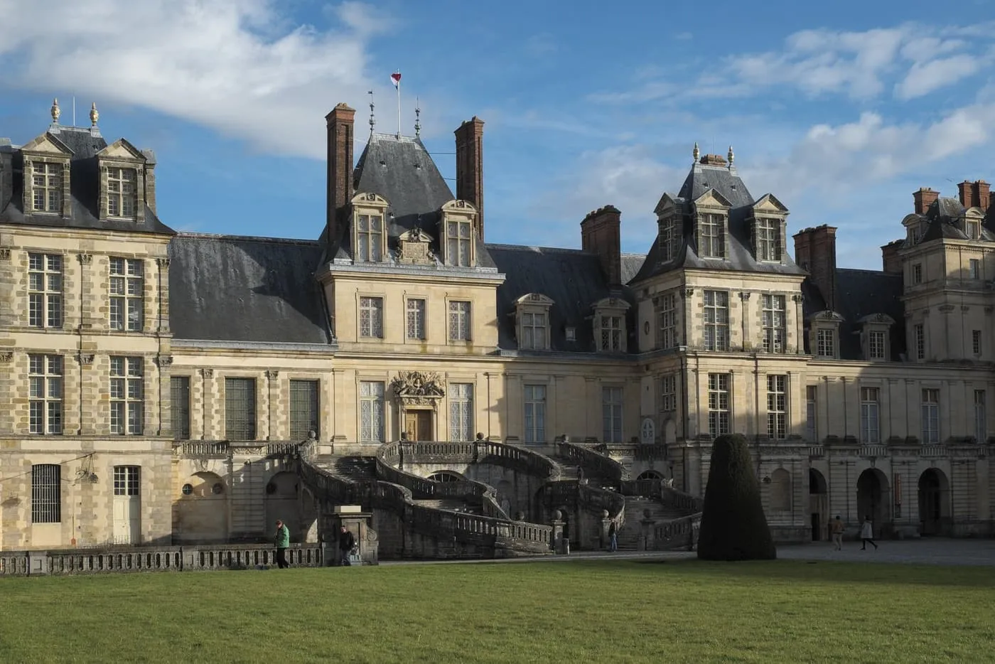 The curving double staircase and slate-roofed wings of the Château de Fontainebleau seen across the lawn of the main courtyard