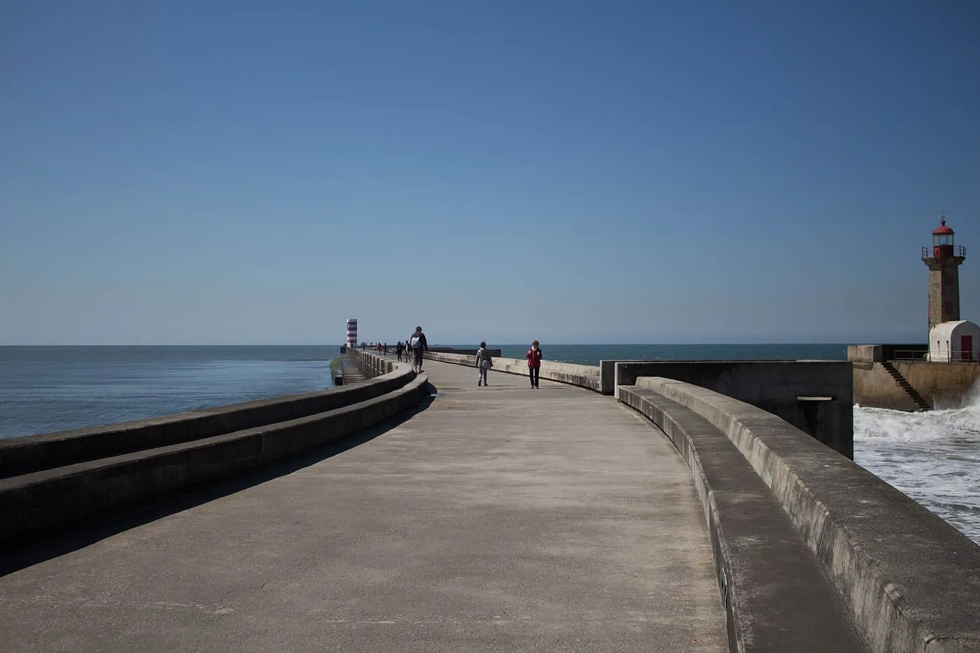 A long concrete pier with a few walkers, a lighthouse at its far end and the sea on both sides under a deep blue sky
