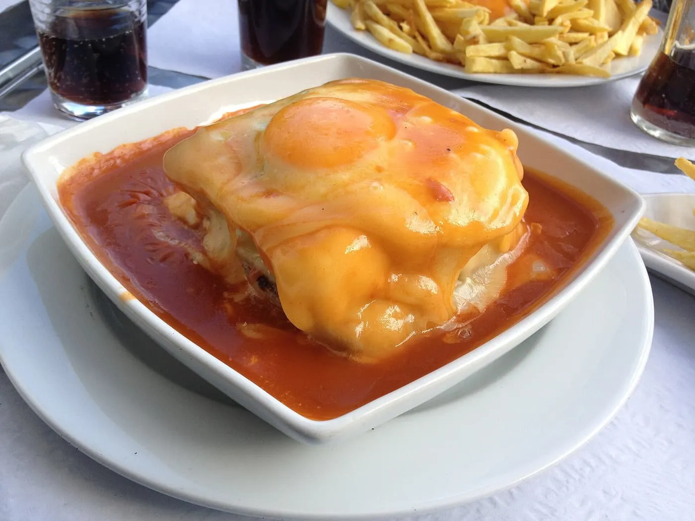 A francesinha in a square white bowl, topped with melted cheese and a fried egg, with chips and drinks on the table