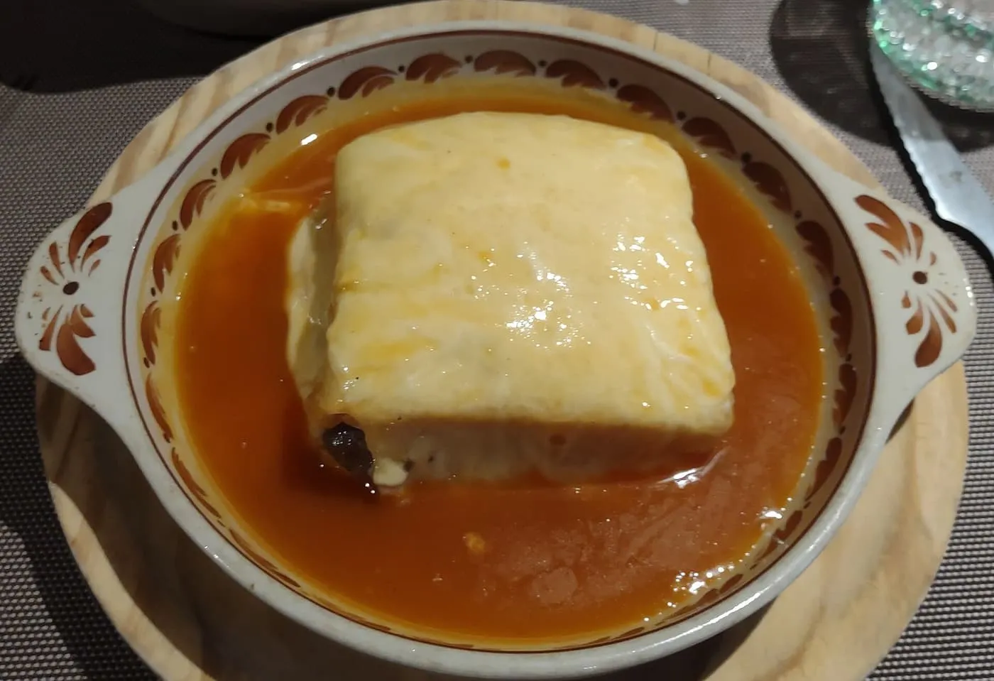 A francesinha in a round dish, a square sandwich under melted cheese sitting in an orange-brown sauce