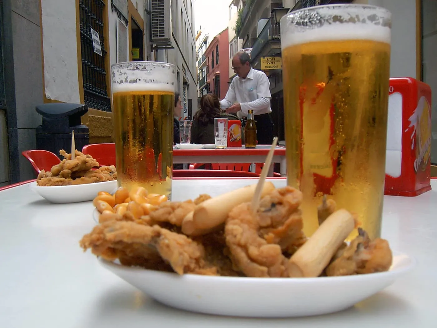 A dish of battered fried anchovies with two glasses of beer on a white street table, and a waiter at work in the lane behind